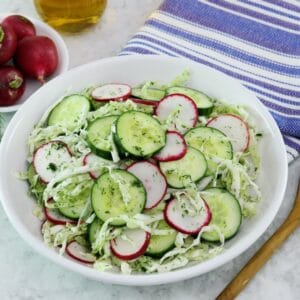 Close up shot of a large bowl filled with crunch pickled salad. A wooden bowl and blue striped napkin sit off to the right side. To the left there is a bunch of fresh radishes.