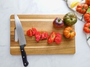 Sliced and chopped heirloom tomatoes with three whole heirloom tomatoes on a cutting board, with a chef's knife resting on the board. Fresh heirloom tomatoes, cloth towel, and lemon slices off to the side.