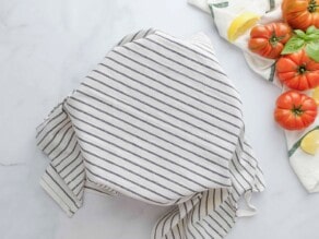 A large bowl covered with a striped cloth towel on a white marble background. Heirloom tomatoes, basil, lemon slices and cloth towel off to the side.