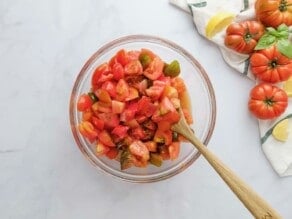 Glass bowl of tomato salad with wooden spoon stirring on a white marble background. Colorful heirloom tomatoes on the side, with a cloth napkin and lemon slices.