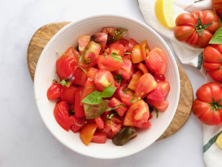 Overhead shot of a white bowl filled with fresh heirloom tomato salad topped with basil. Bowl rests on a wooden cutting board on a white marble background, with large fresh heirloom tomatoes, a linen towel, and a slice of lemon on the side.