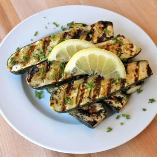 Grilled Lemon Butter Zucchini served on a white plate with 3 slices of lemon and chopped herbs