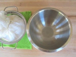 Stainless steel mixing bowl, cheesecloth yogurt bundle in mesh strainer.