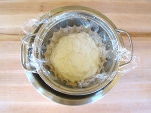 Coffee filter-lined mesh colander with yogurt, covered by plastic wrap.
