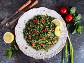 Overhead shot of quinoa tabbouleh salad in a white dish with lemon slices, half lemon, mint sprigs, green onions and small tomatoes. Salad fork and spoon on the upper left of the dish.