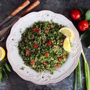 Overhead shot of quinoa tabbouleh salad in a white dish with lemon slices, half lemon, mint sprigs, green onions and small tomatoes. Salad fork and spoon on the upper left of the dish.
