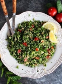 Overhead shot of quinoa tabbouleh salad in a white dish with lemon slices, half lemon, mint sprigs, green onions and small tomatoes. Salad serving utensils with wooden handles rest in the salad.