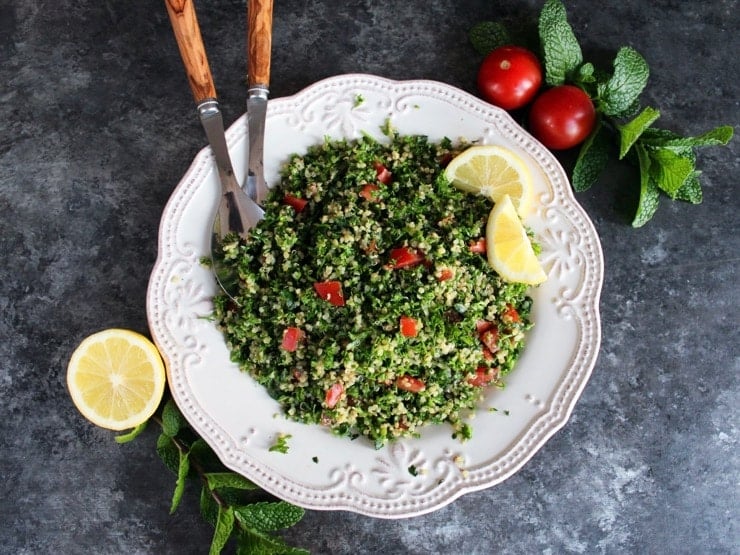 Overhead shot of quinoa tabbouleh salad in a white dish with lemon slices, half lemon, mint sprigs, green onions and small tomatoes. Salad serving utensils with wooden handles rest in the salad.