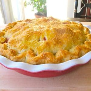 Golden baked pie in red pie dish on wooden cutting board on windowsill.