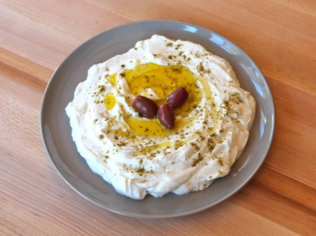 Plato de yogur colado labneh sobre tabla de cortar de madera, cubierto con aceite de oliva, za'atar y aceitunas.'atar and olives.