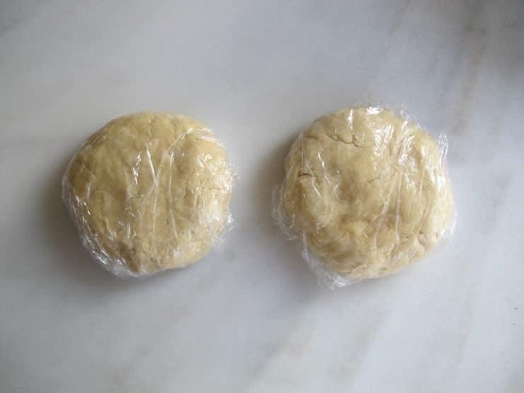 Two disks of pie crust dough wrapped in plastic on marble board.
