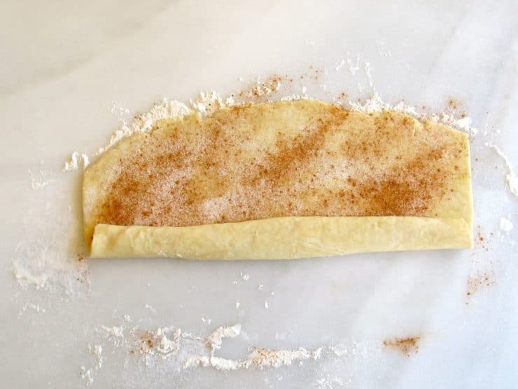 Rolling pie crust pinwheel dough filled with sugar on marble surface.