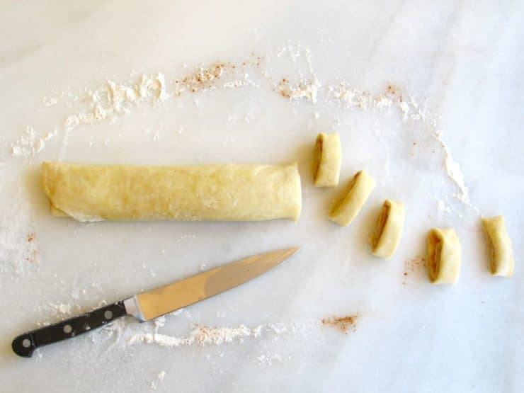 Slicing pie crust pinwheel rolled dough with knife on marble surface.