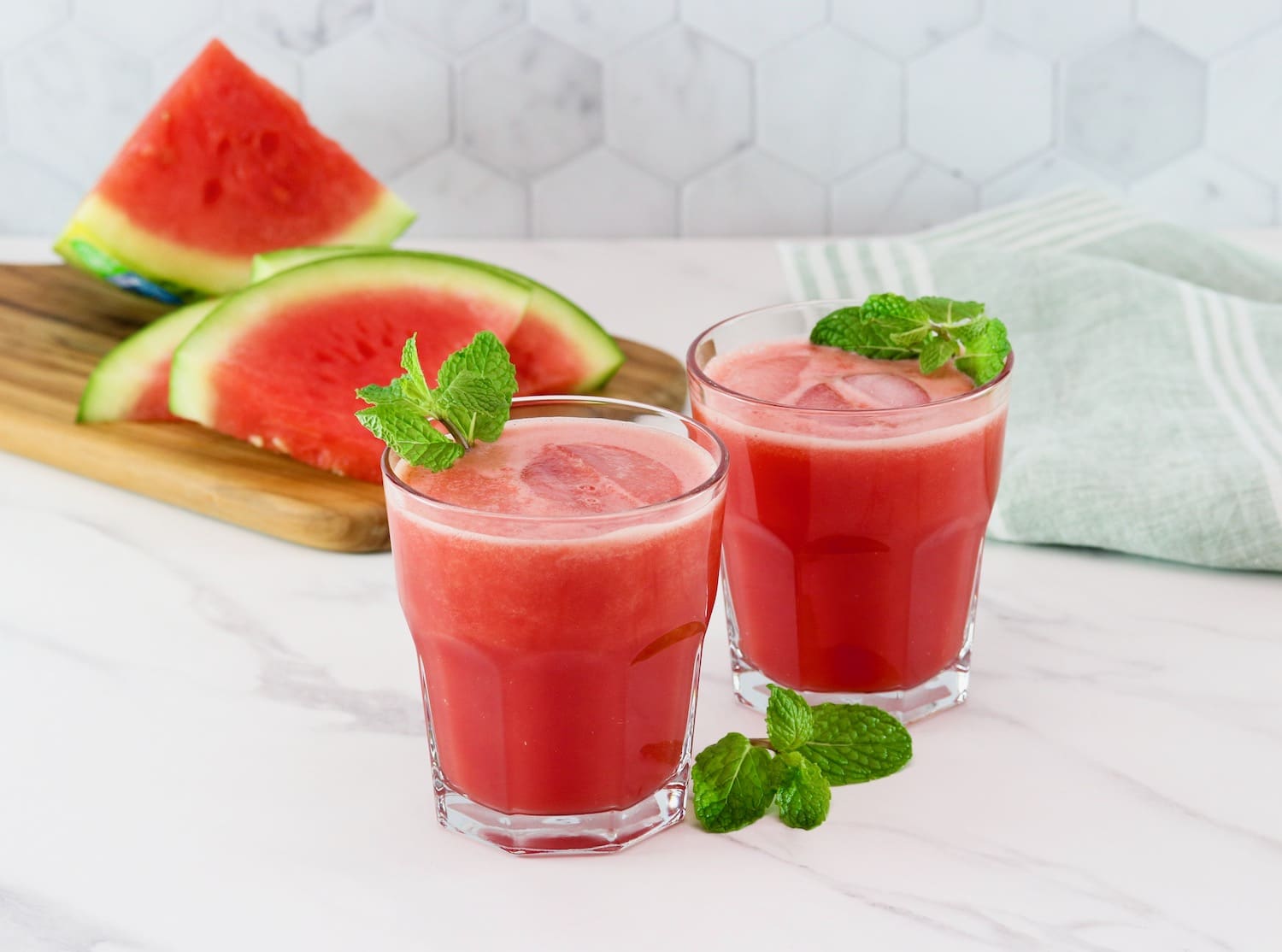 Horizontal shot of two short glasses filled with watermelon rum cocktail, garnished with fresh mint. Slices of watermelon sit on top of a wooden cutting board on the left side.