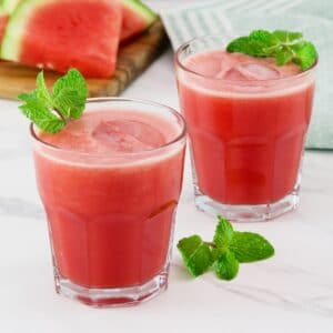 Horizontal shot of two short glasses filled with a frozen watermelon rum cocktail, garnished with fresh mint. Slices of watermelon sit on top of a wooden cutting board on the left side.