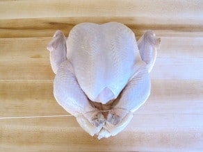 Whole chicken on cutting board - trussing strings looped around drumsticks, pulling drumsticks tight together.