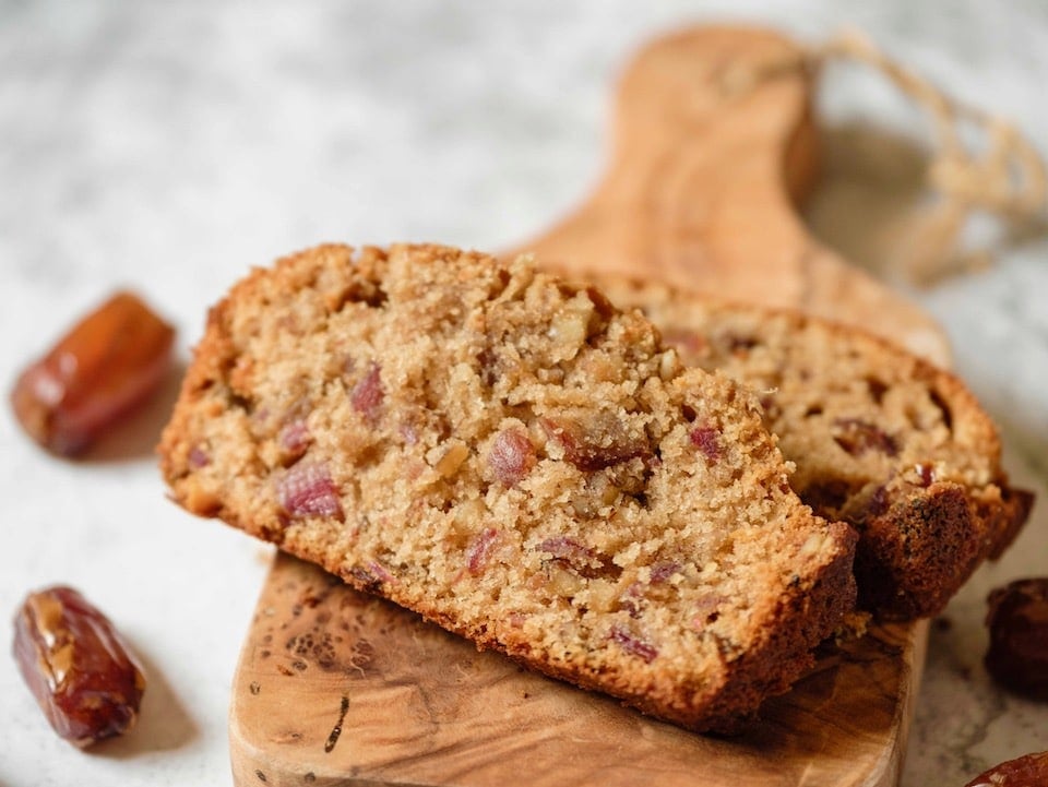 Two slices of moist, crumby date honey nut cake on a wooden cutting board on marble background, two dates resting beside it on the counter.