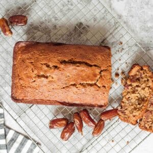 Wide horizontal shot - sliced date honey nut cake on cooling rack with luscious dates on marble countertop, striped linen towel laying beside the rack.