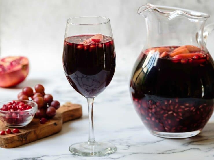 Horizontal shot - Glass of dark red Rosh Hashanah Sangria with a pitcher of sangria in background, each with fruit pieces floating on top. Wood cutting board with grapes and pomegranate arils, half a pomegranate in background.