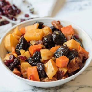 Horizontal shot - white bowl of tzimmes with sweet potatoes, prunes, carrots, and dried cranberries on a marble countertop, linen napkin with dried cranberries in background.