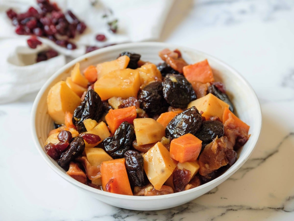 Horizontal shot - white bowl of tzimmes with sweet potatoes, prunes, carrots, and dried cranberries on a marble countertop, linen napkin with dried cranberries in background.