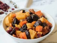 Square featured shot - white bowl of tzimmes with sweet potatoes, prunes, carrots, and dried cranberries on a marble countertop, linen napkin with dried cranberries in background.