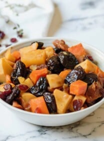 Square featured shot - white bowl of tzimmes with sweet potatoes, prunes, carrots, and dried cranberries on a marble countertop, linen napkin with dried cranberries in background.