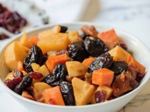 Close up over rim of bowl - white bowl of tzimmes with sweet potatoes, prunes, carrots, and dried cranberries on a marble countertop, linen napkin with dried cranberries in background.