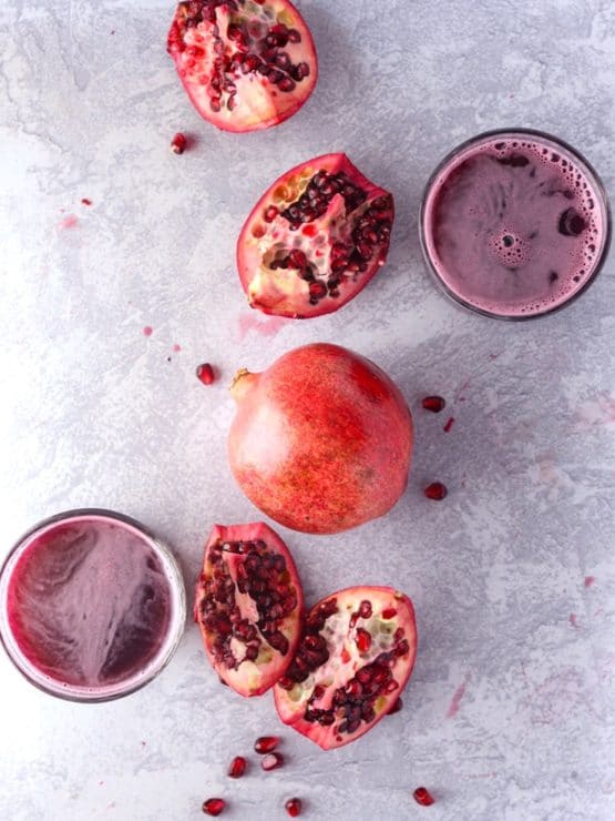 Whole pomegranate and pomegranate slices with scattered seeds and two glasses of juice on a marble background.