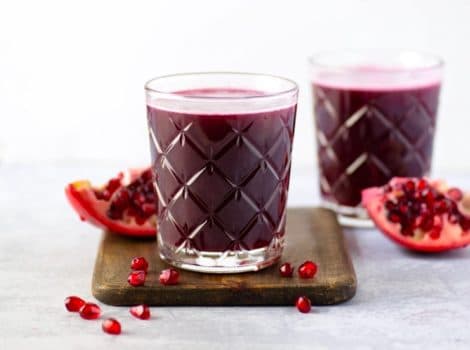 Two glasses of pomegranate juice surrounded by pomegranate slices and seeds on a wooden cutting board with marble background.