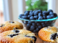 Freshly baked Buttermilk Blueberry Muffins on a cutting board with a side of fresh blueberries in a bowl