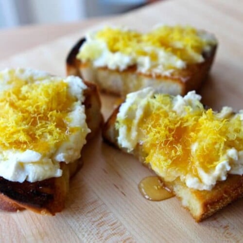 Three slices of Challah Bruschetta with lemon ricotta drizzled with honey on a cutting board