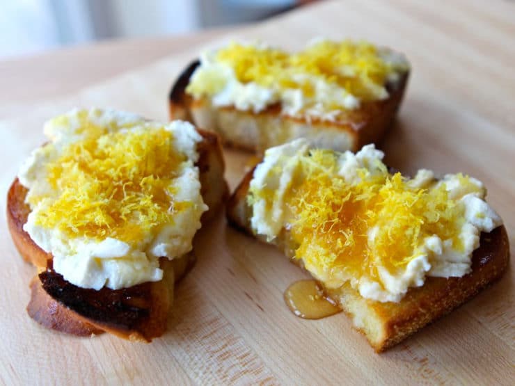 Three slices of Challah Bruschetta with lemon ricotta drizzled with honey on a cutting board