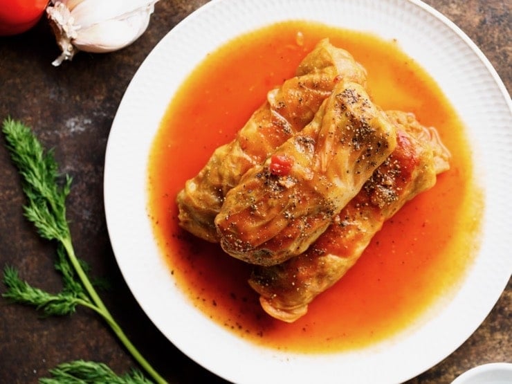 Three stuffed cabbage leaves - holishkes on a plate with sauce, fresh dill and garlic on the side.