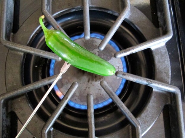 Fresh jalapeño held by metal skewer fork over gas flame on stovetop.