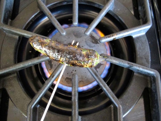 Charred jalapeño held by metal skewer fork over gas flame on stovetop.