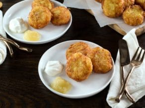 Horizontal image - three golden potato latkes on a white plate with sour cream and applesauce, fork and knife with cloth napkin beside. More latkes on plates in backgrounds.