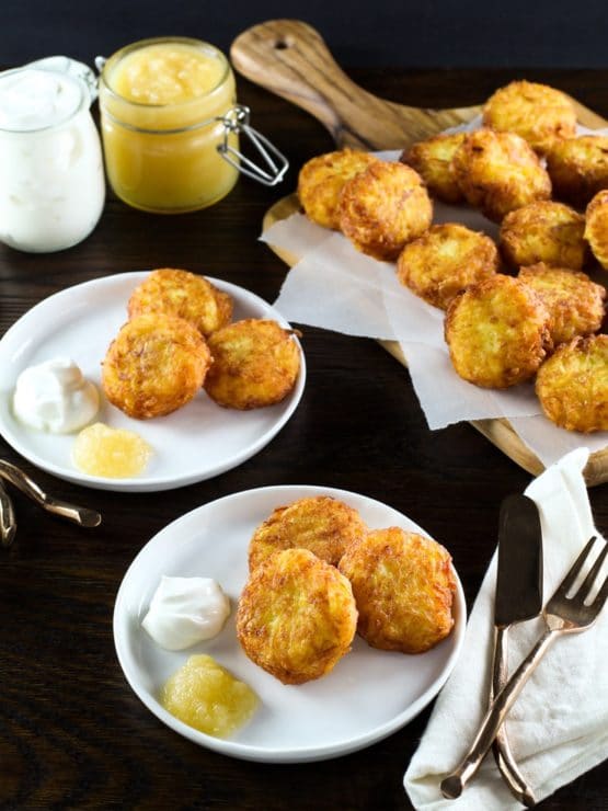 Vertical shot - crisp latkes on white plates with applause and sour cream. Pile of latkes on a cutting board in background.