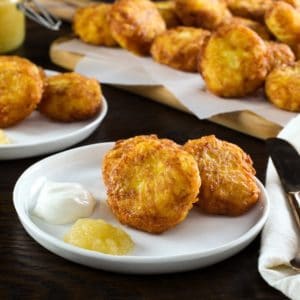 Close up shot - crispy golden latkes on a white plate with sour cream and applesauce. Fork and knife with cloth napkin on the side. Pile of potato pancakes in the background.