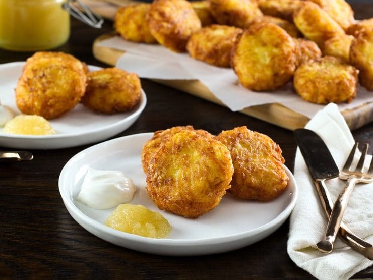 Close up shot - crispy golden latkes on a white plate with sour cream and applesauce. Fork and knife with cloth napkin on the side. Pile of potato pancakes in the background.