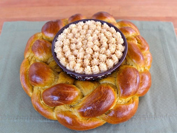 Horizontal close-up of a pumpkin challah centerpiece with honey butter, resting on a green checkered table napkin.