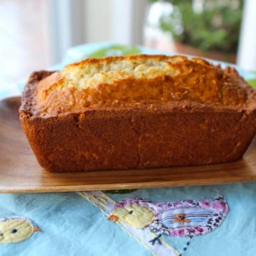 A golden-brown coconut cake sits on a brown, upward-curved plate, rests on a tablecloth adorned with a charming chick design.