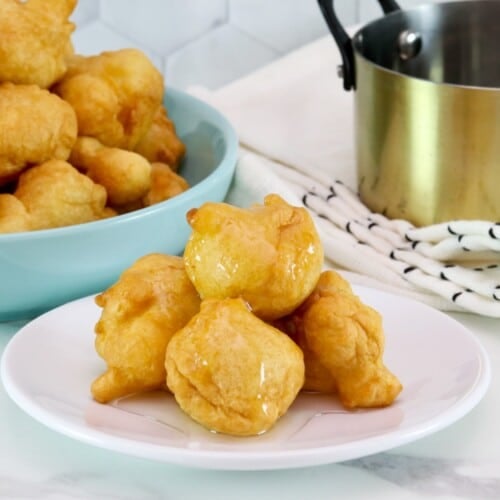 Close up - Hanukkah buñuelos dessert - pile of four on a small white plate drenched in clear syrup. Large plate of buñuelos in background, as well as a gold pot of clear sauce, on a white and blue linen towel.