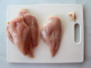 4 chicken breasts on a white cutting board, one breast trimmed of excess fat.