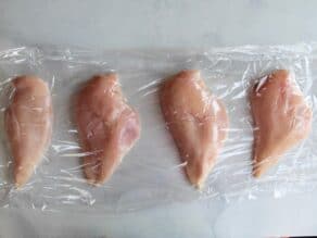 4 chicken breasts sandwiched between two layers of plastic wrap on countertop, spaced a couple of inches apart.
