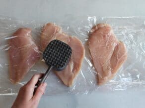 Hand pounding out chicken breasts between layers of plastic wrap with a metal mallet.
