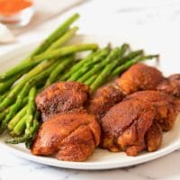 Horizontal Crop - Smoked Paprika Chicken Thighs with boneless skinless thighs, roasted asparagus in background, on a white plate on a white marble countertop, dish of smoked paprika and garlic cloves in background.