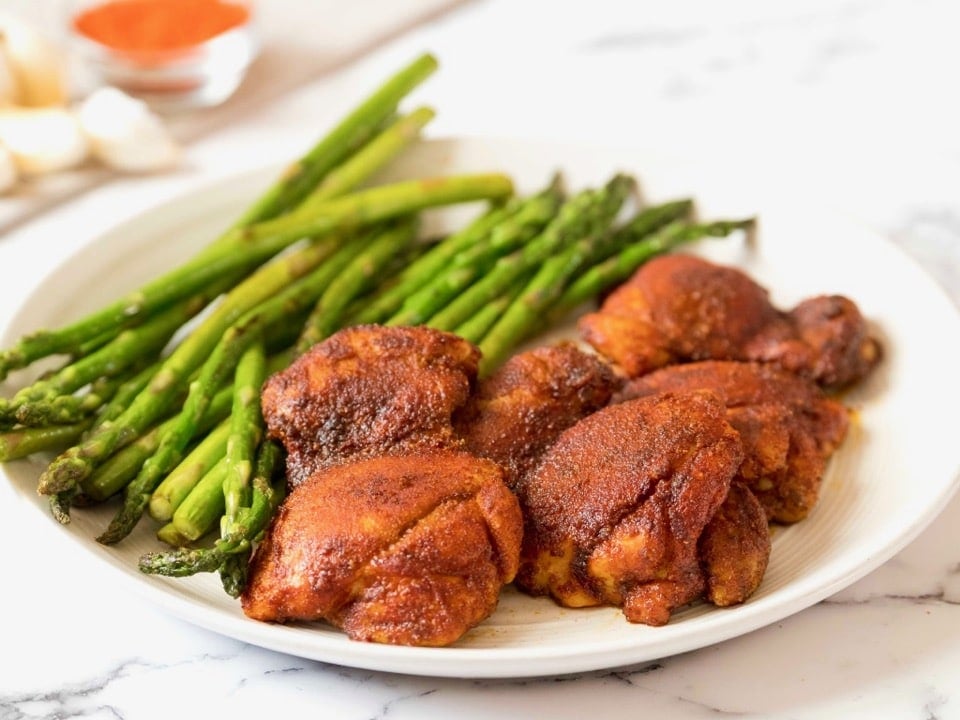 Horizontal Crop - Smoked Paprika Chicken Thighs with boneless skinless thighs, roasted asparagus in background, on a white plate on a white marble countertop, dish of smoked paprika and garlic cloves in background.