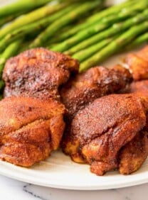 Square Crop - Smoked Paprika Chicken Thighs with boneless skinless thighs, roasted asparagus in background, on a white plate on a white marble countertop.