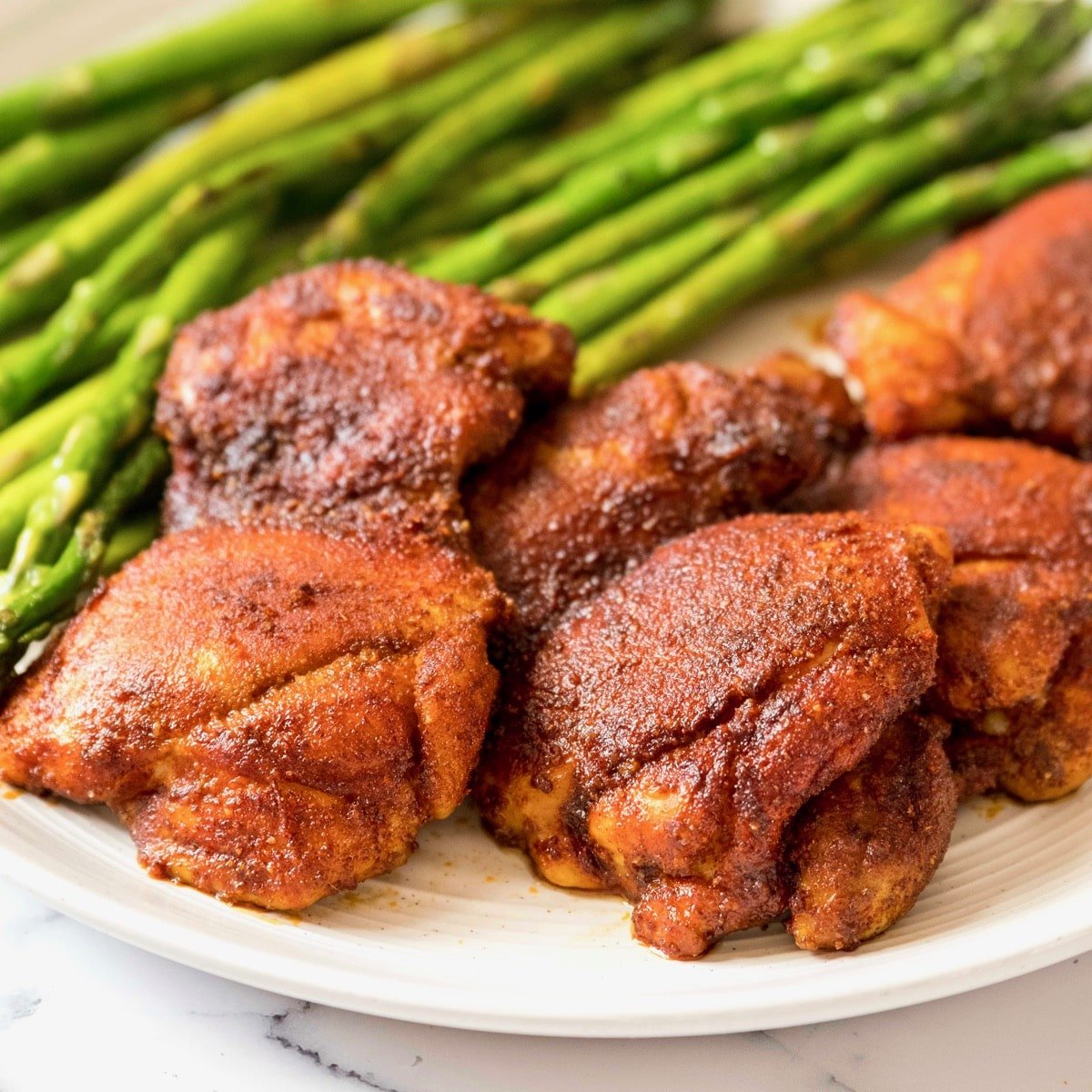 Square Crop - Smoked Paprika Chicken Thighs with boneless skinless thighs, roasted asparagus in background, on a white plate on a white marble countertop.